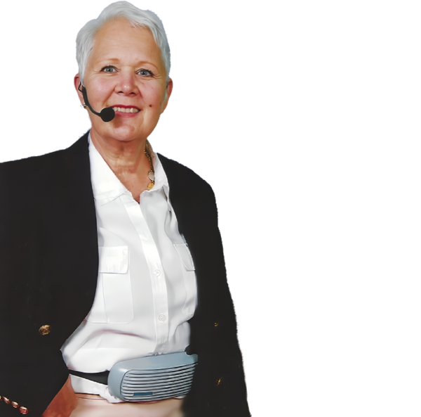 An older woman with short gray hair wears a ChatterVOX® Pro w/headset mic and a black blazer over a white shirt, smiling as she stands against a plain white background, with the device secured around her waist.
