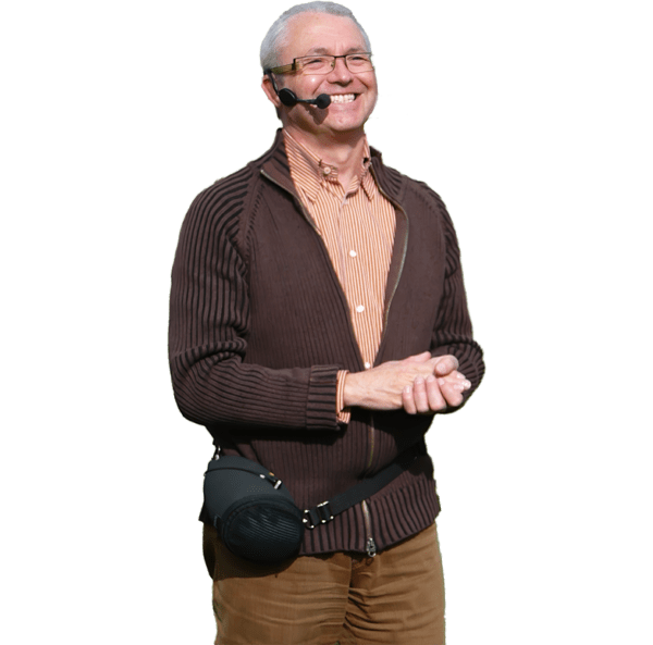 An older man with gray hair and glasses smiles while wearing a ChatterVOX® Model 6 Coaching Amplifier headset and speaker around his waist, dressed in a brown zip-up sweater over a striped shirt.