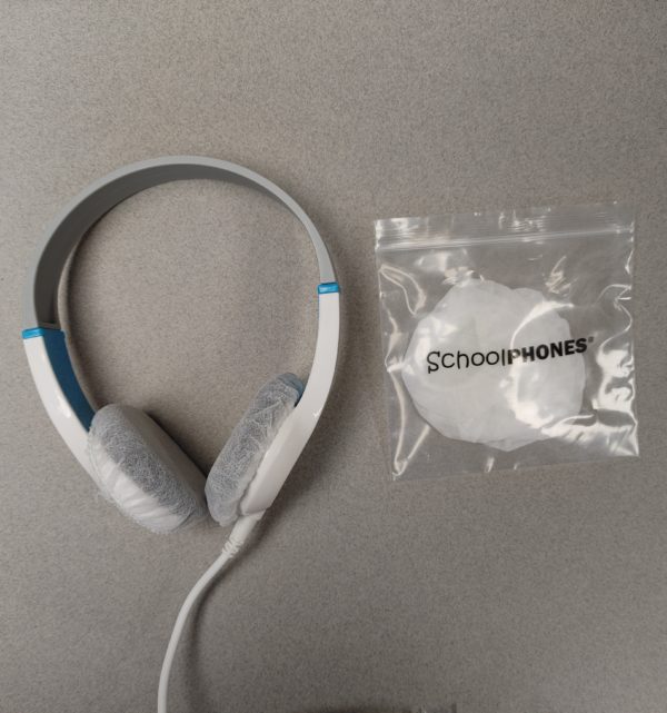 A pair of white school headphones with K6 / BK Series - Hygienic/Sanitary covers (250 Pairs) on the ear pads sits next to a small bag labeled SchoolPHONES on a gray surface.